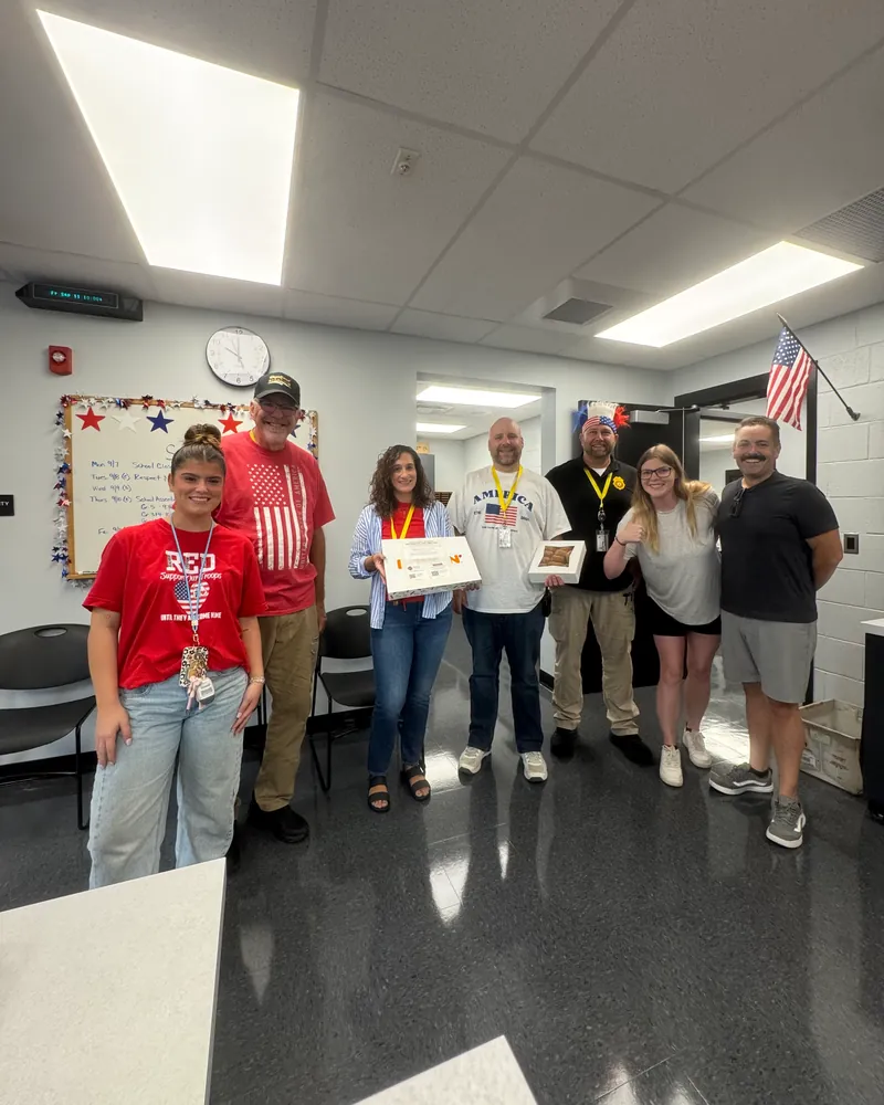 Eric and a teammate with school staff in a staff room decorated with stars and a flag, holding boxes of treats